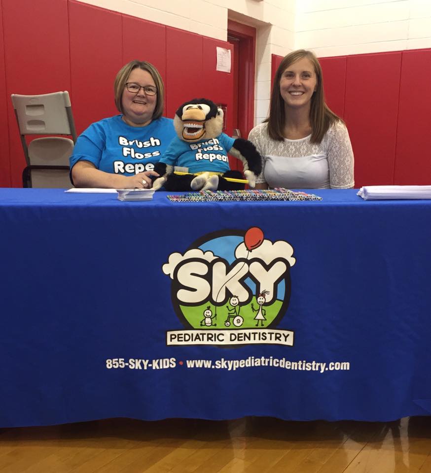 Two women at Sky Pediatric Dentistry booth smiling behind blue table with red wall backdrop.