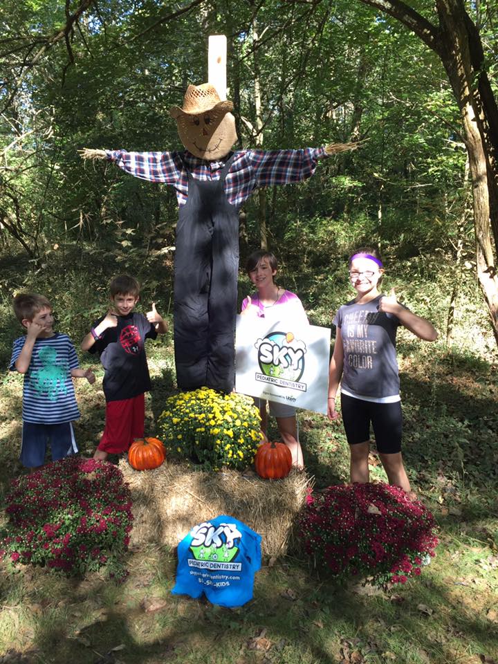 Children gathered around scarecrow decoration in wooded outdoor setting during fall celebration.