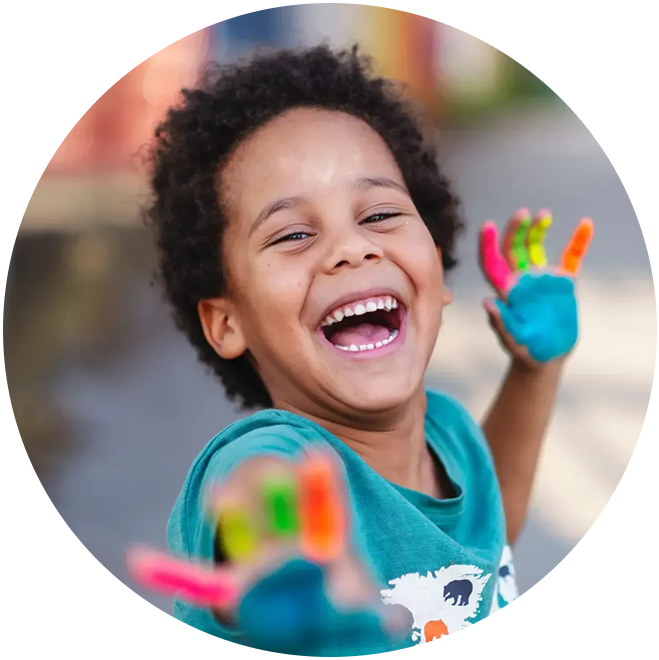 Joyful child with painted hands laughing, wearing blue shirt in bright outdoor setting.