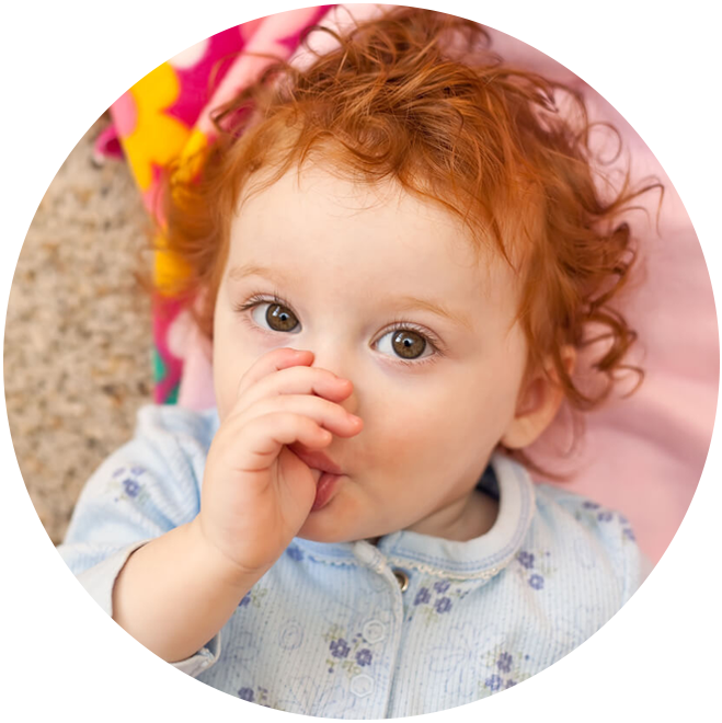 Toddler with curly red hair sucking thumb against pink background with colorful bow.