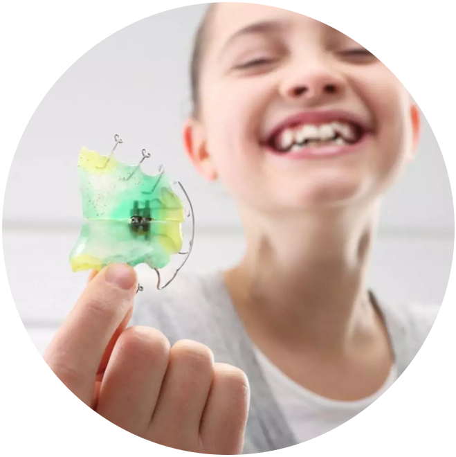 Smiling child holding green orthodontic retainer or dental appliance in dental office setting.