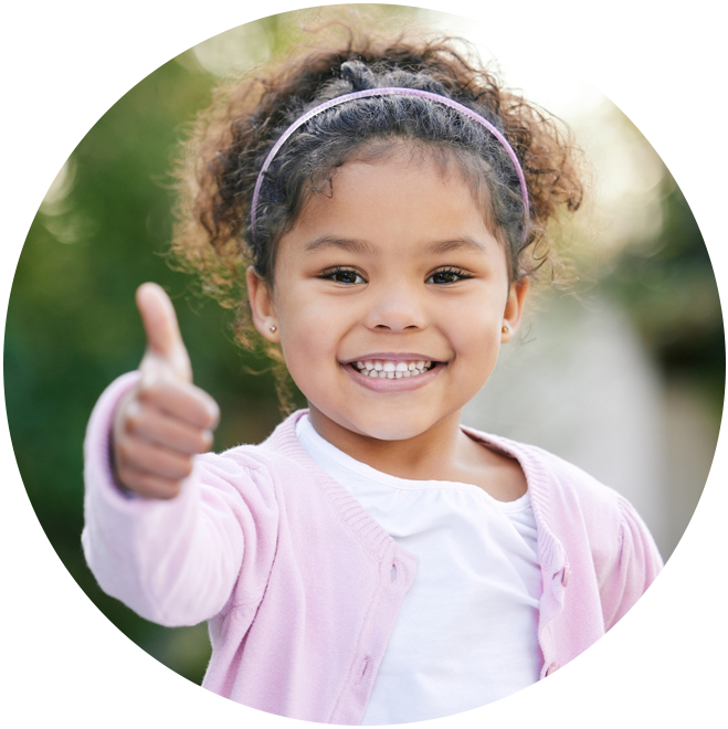 Young child in pink cardigan giving thumbs up outdoors, smiling at camera.