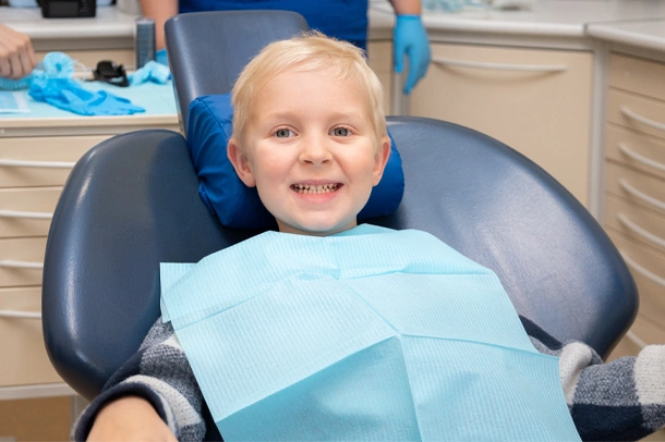 Young child smiling in dental chair wearing blue bib at pediatric dentistry office.
