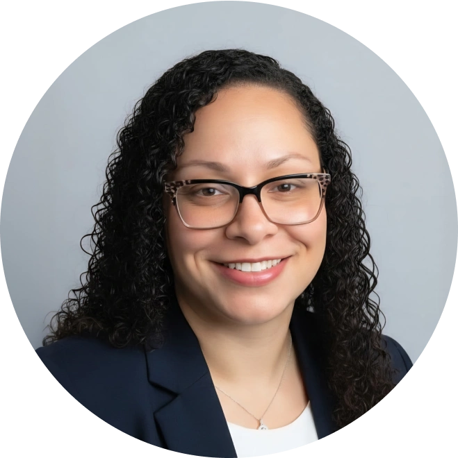 Professional headshot of a woman with long dark curly hair and glasses, smiling at camera in business attire.